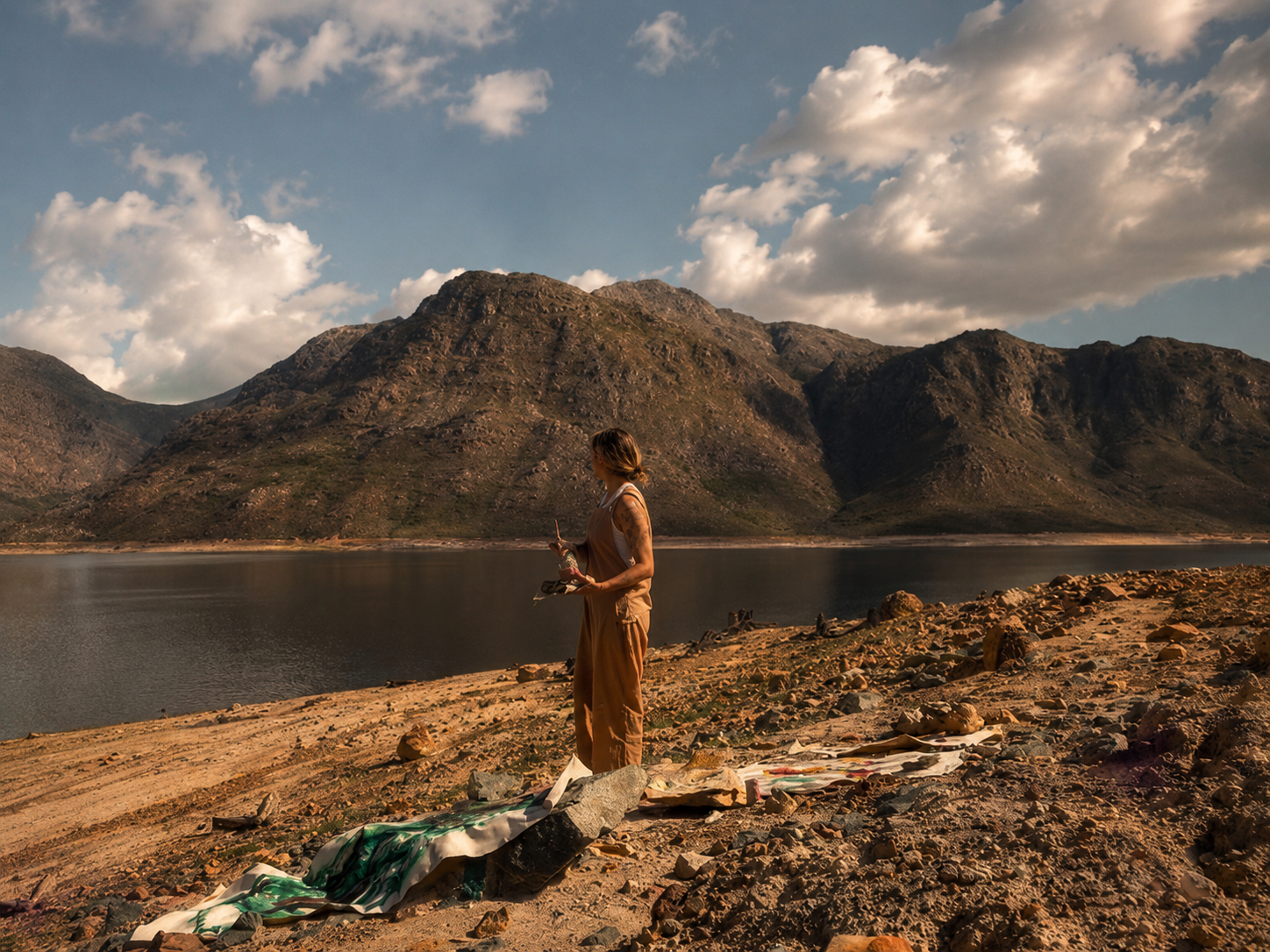Nicky Myny painting outdoors by a South African mountain lake, unstretched canvases laid on the shore